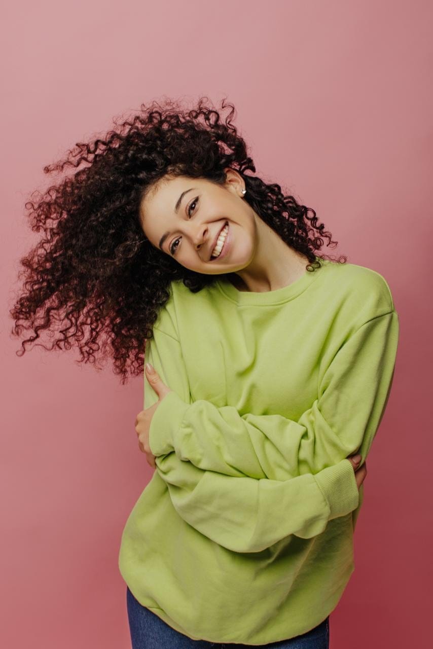 A smiling young woman with curly hair hugs herself in a soft green sweatshirt.